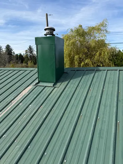 Green metal chimney encasement with a round cap and base flashing on a green metal roof in Campbell River.