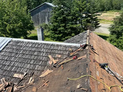 Cedar shakes being removed from intersecting roof slopes, with exposed decking, loose shakes and tools visible.