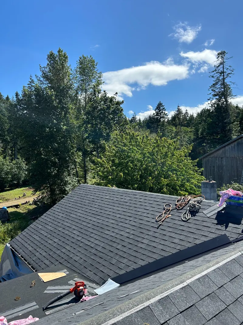 New shingles across intersecting roof slopes beside a chimney, with tools, ropes and an unfinished section in the foreground.