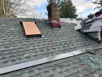 Shingle installation in progress around a brick chimney and a temporarily covered roof opening, with metal flashing pieces nearby.