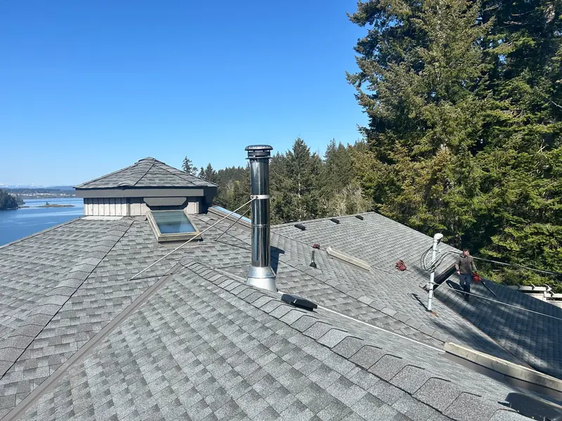 Grey shingle roof with multiple hips and valleys, a skylight and a metal chimney, with water and trees beyond.