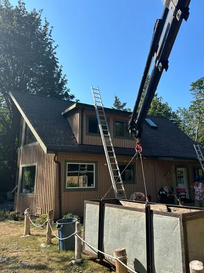 Dark shingles around a wide two-window dormer and a skylight on a timber-sided home, with ladders and lifting equipment still on site.