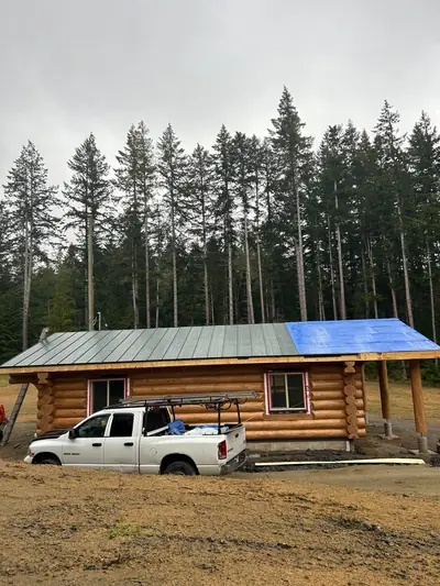 Log building with a partially installed grey metal roof and blue underlayment at one end, surrounded by trees.
