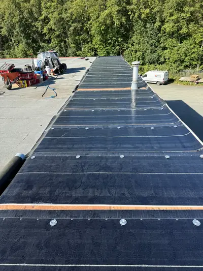 Dark roofing membrane being laid across a low-slope roof, with fasteners, a vent and tools visible.