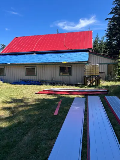 New red metal panels on an upper roof, with blue underlayment on the lower slope and panels staged on the lawn.