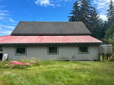Weathered shingles on an upper roof above a faded red metal lower roof, with three green-trimmed windows below.