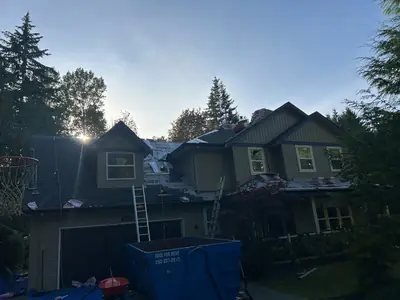 Multi-gable residential roof with shingles and underlayment visible, materials staged above and a disposal bin below.