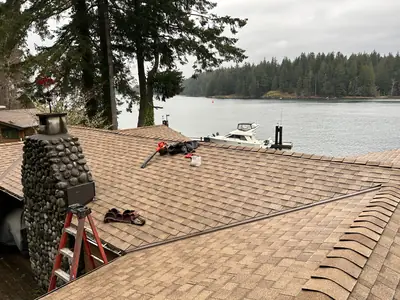 Brown shingle roof beside the water with a stone chimney, metal valley and tools on the roof.
